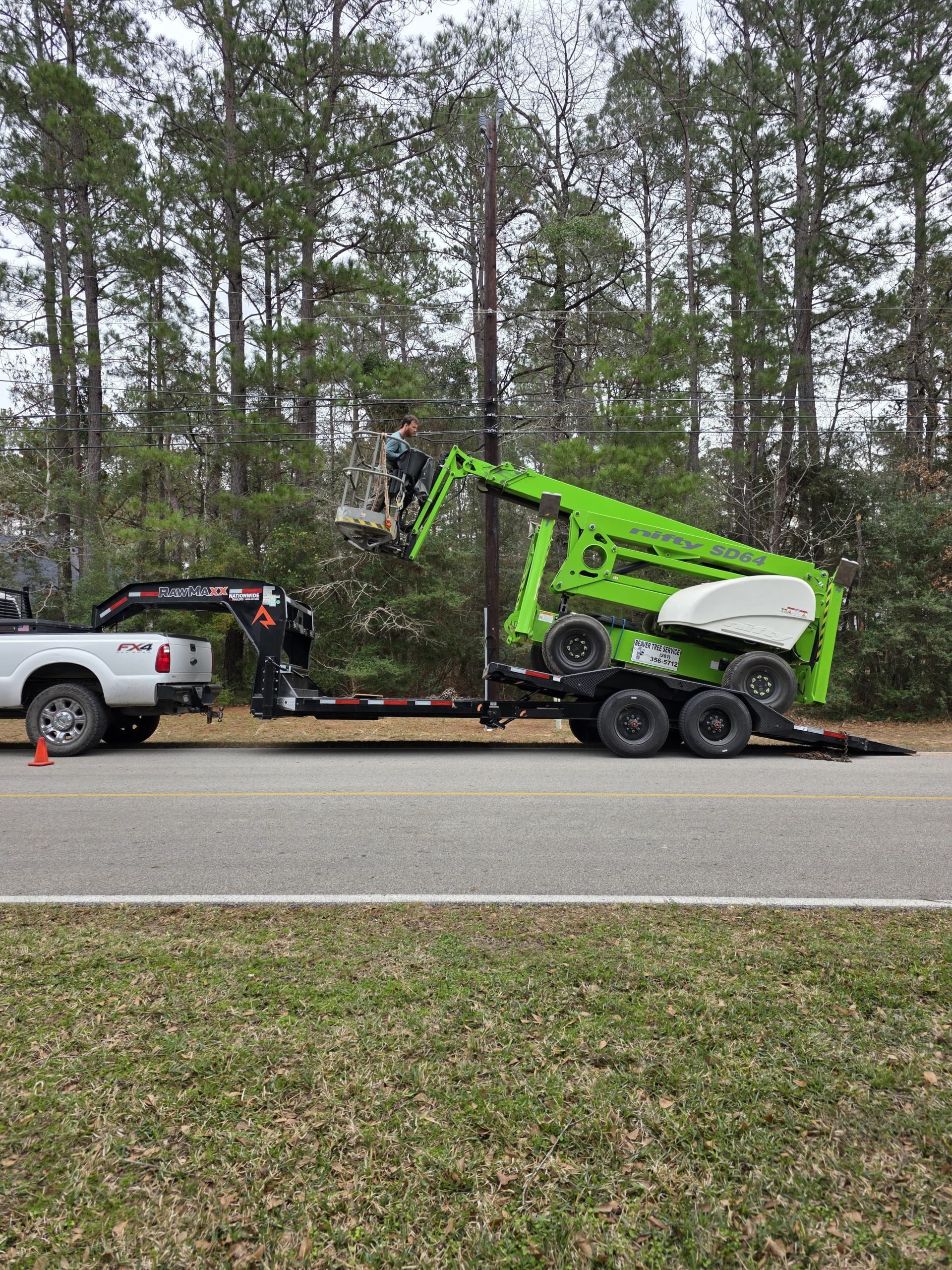 A green tree service lift truck parked on a road near a wooded lot.